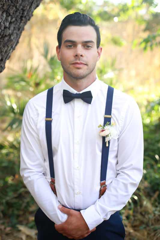 Man wearing a white shirt, black bow tie, and navy suspenders standing outdoors with greenery in the background.