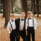 Man in a suit with two boys in formal attire standing in a forest. black bow ties and suspenders