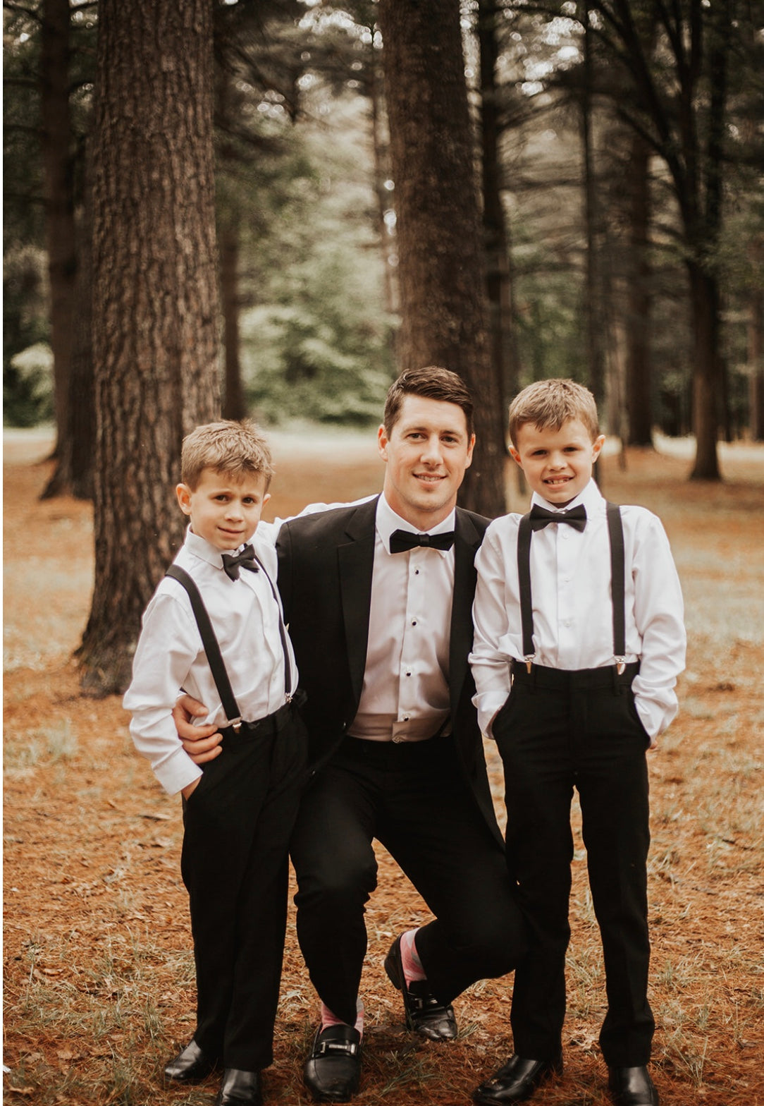 Man in a suit with two boys in formal attire standing in a forest. black bow ties and suspenders