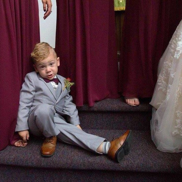 Young boy in a suit sitting on steps with wine cabernet bridesmaids and a wedding dress in the background. Wine bow tie