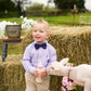 Child in a checkered shirt and Plum bow tie feeding a lamb with flowers on a farm setting.