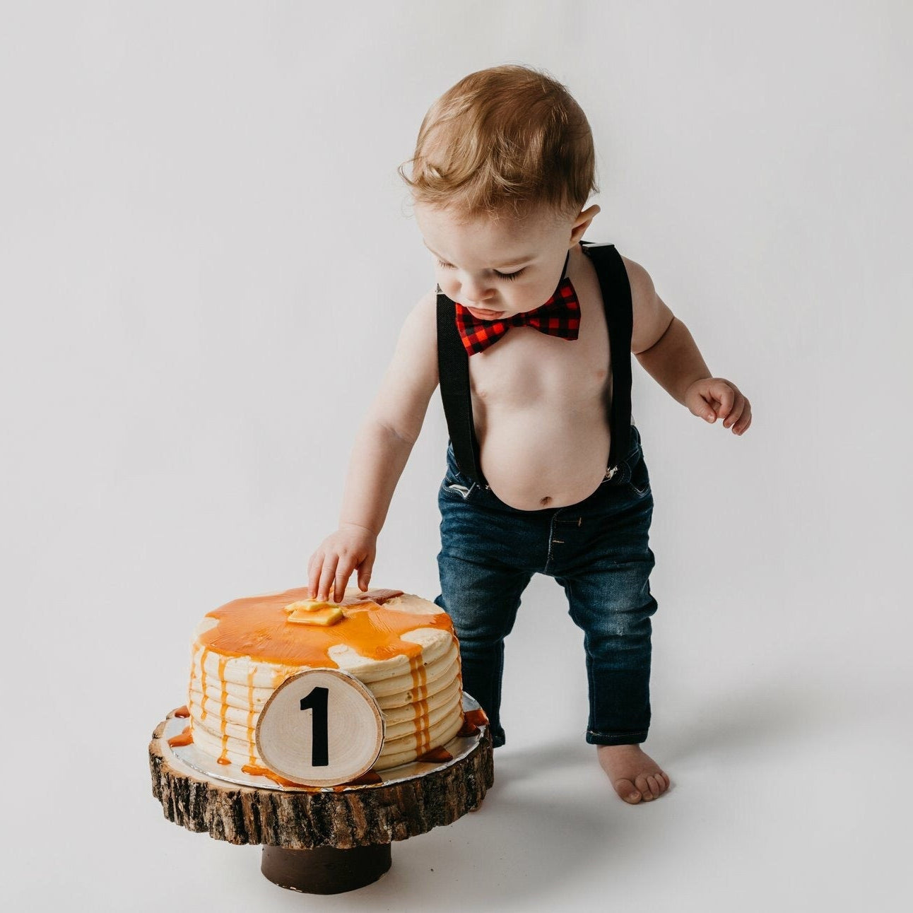 Child in suspenders standing next to a small cake with a number 1 on a white background