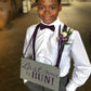 Young boy in formal attire Plum bow tie and suspenders holding a "last chance to run" sign with text in a church