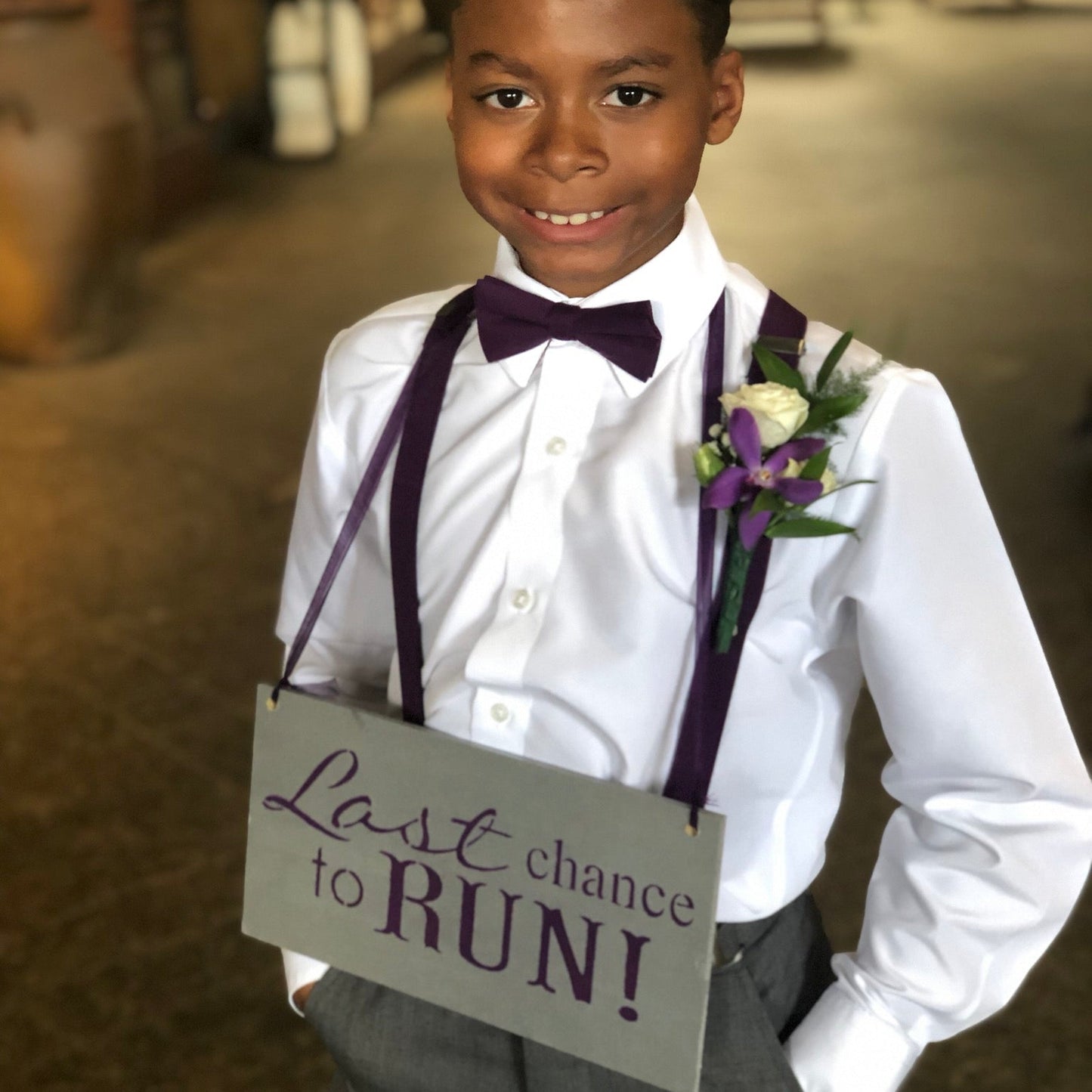 Young boy in formal attire Plum bow tie and suspenders holding a "last chance to run" sign with text in a church