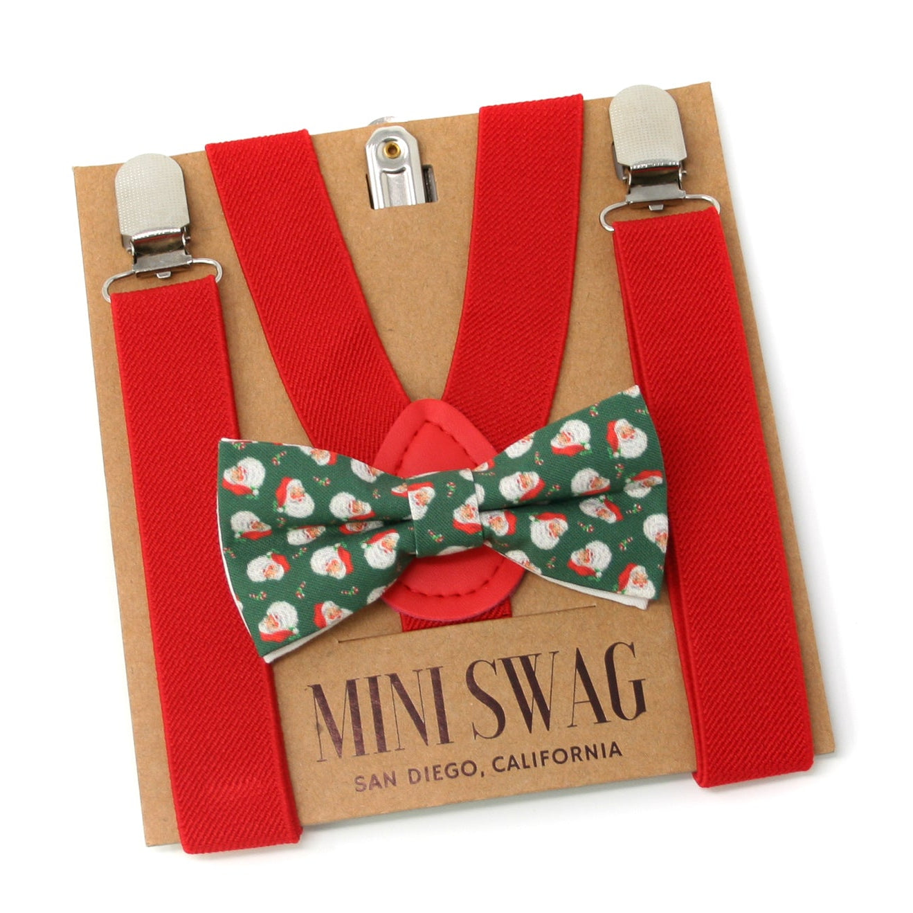 Red suspenders with a green bow tie featuring Santa Claus patterns on a cardboard backing with 'Mini Swag' branding.