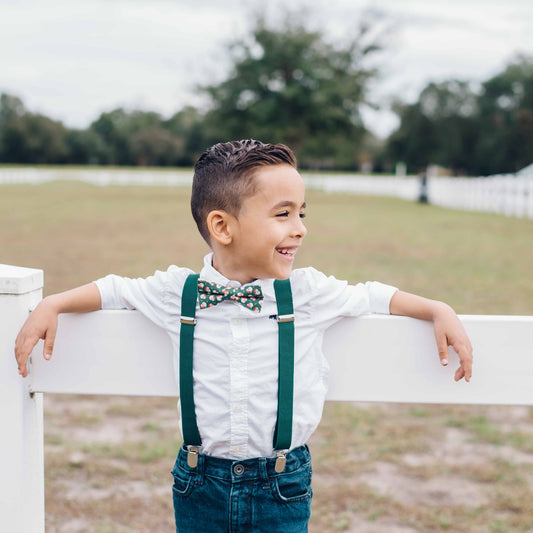 Child wearing suspenders and a bow tie standing on a white fence in an open field.
