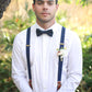 Man wearing a white shirt, black bow tie, and navy suspenders standing outdoors with greenery in the background.