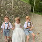 Ring bearers wearing sea glass bow ties and camel leather buckle suspenders on a swing set with a stone wall and greenery in the background