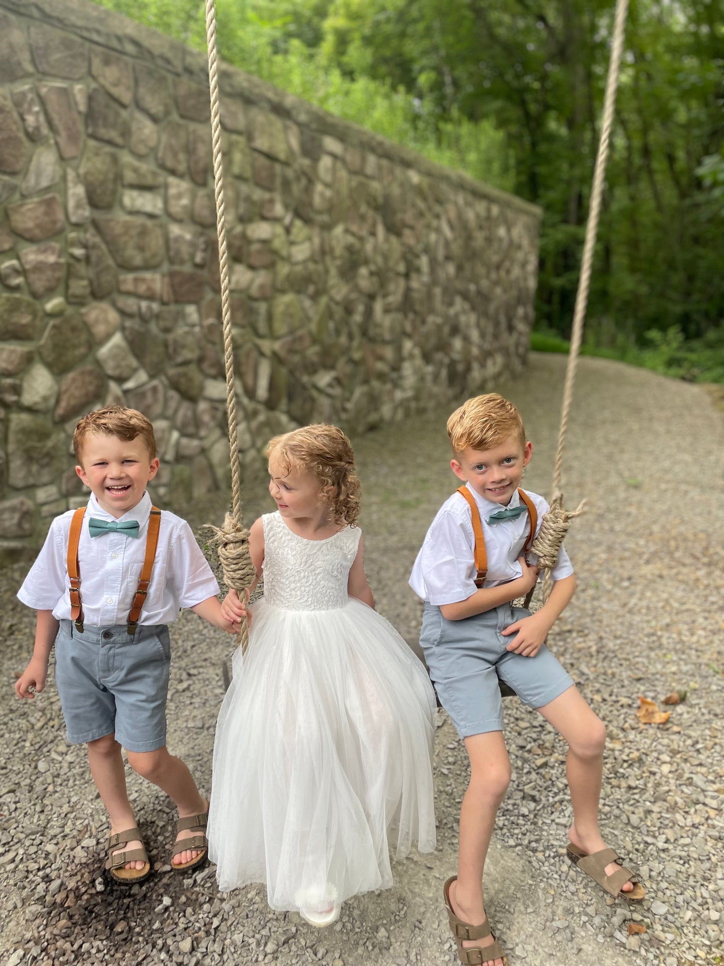 Ring bearers wearing sea glass bow ties and camel leather buckle suspenders on a swing set with a stone wall and greenery in the background