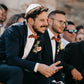 Two men in suits with bow ties sitting outdoors, one wearing a yarmulke.