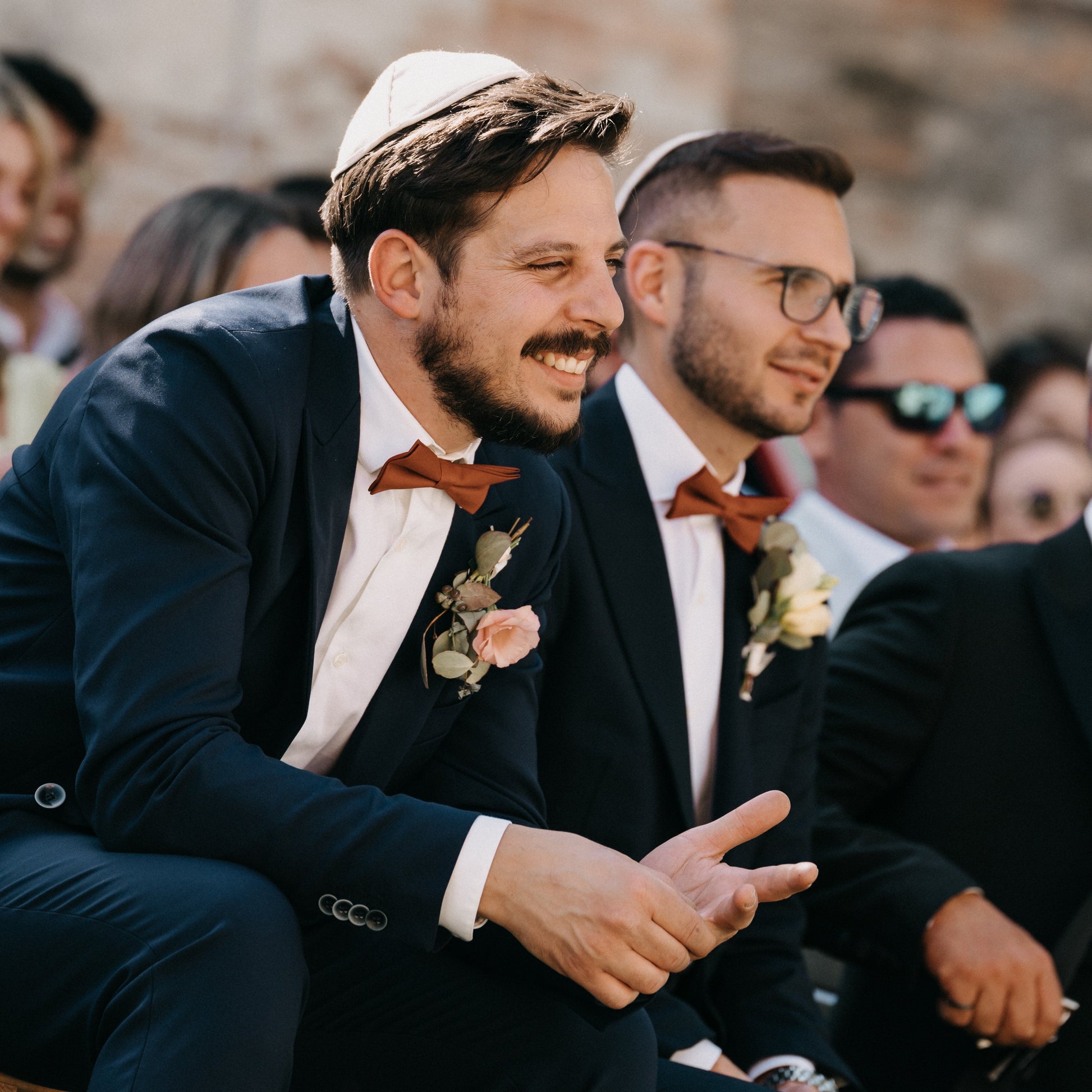 Two men in suits with bow ties sitting outdoors, one wearing a yarmulke.