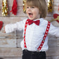 Child wearing a white shirt with a red bow tie and suspenders, standing against a wooden wall with heart decorations.
