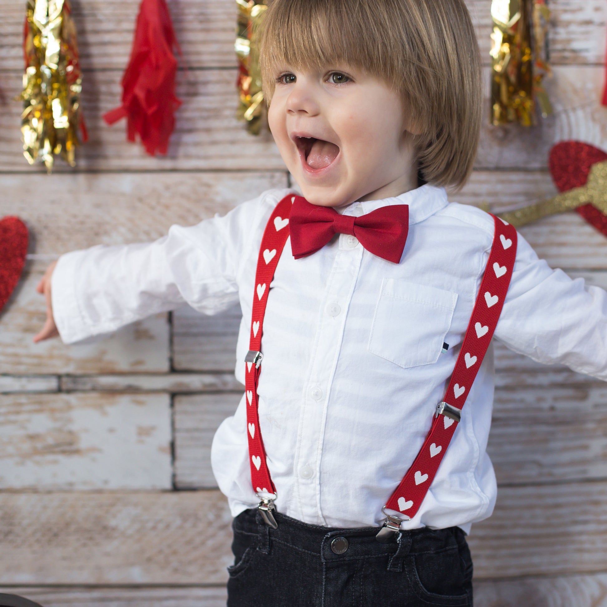 Child wearing a white shirt with a red bow tie and suspenders, standing against a wooden wall with heart decorations.