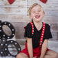 Child sitting on a white rug with red heart suspenders and a wooden backdrop with hearts and arrows. Wearing heart suspenders