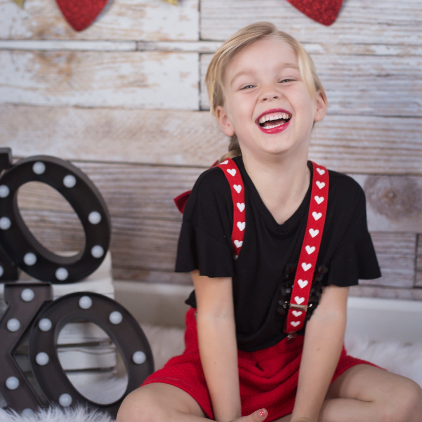 Child sitting on a white rug with red heart suspenders and a wooden backdrop with hearts and arrows. Wearing heart suspenders