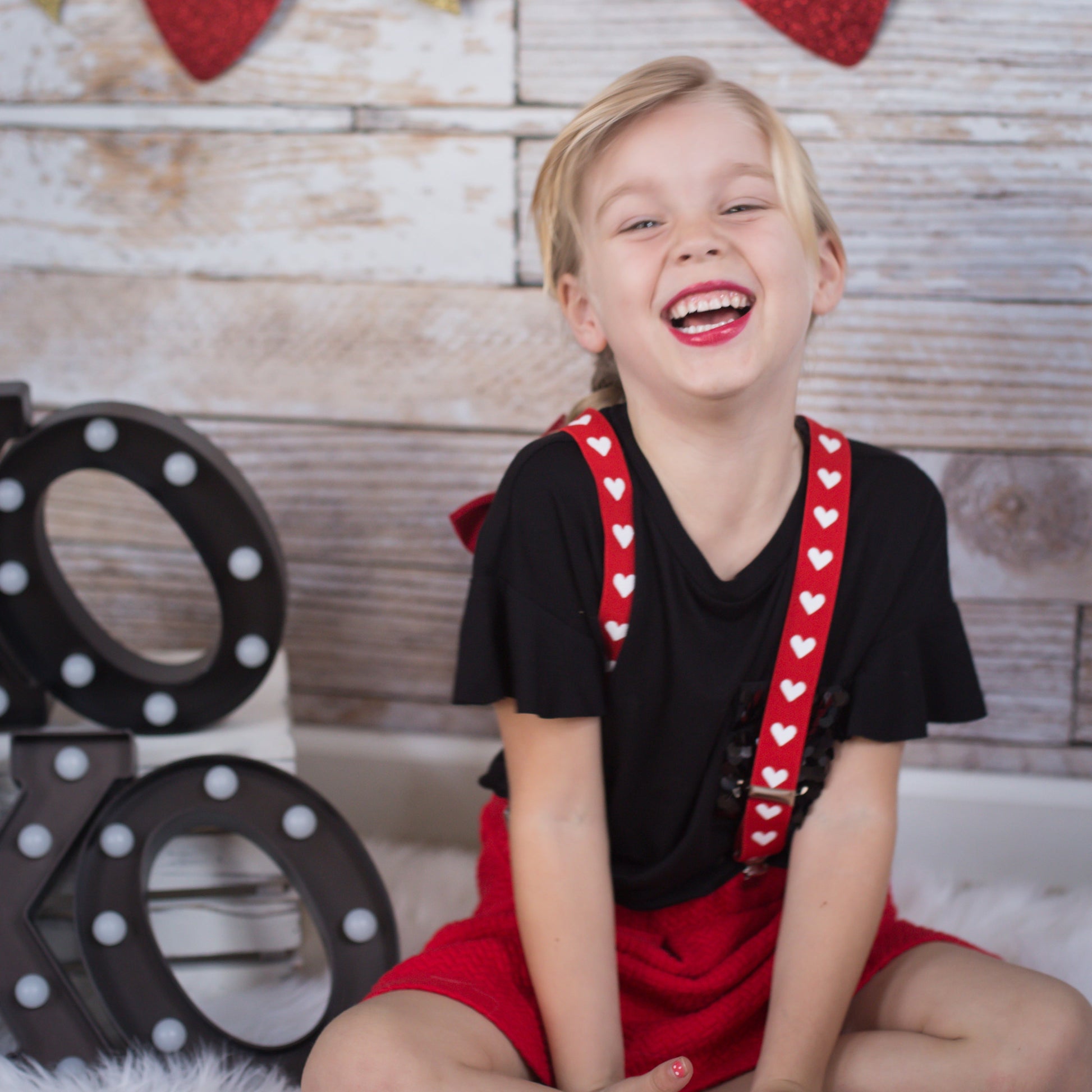Child sitting on a white rug with red heart suspenders and a wooden backdrop with hearts and arrows. Wearing heart suspenders