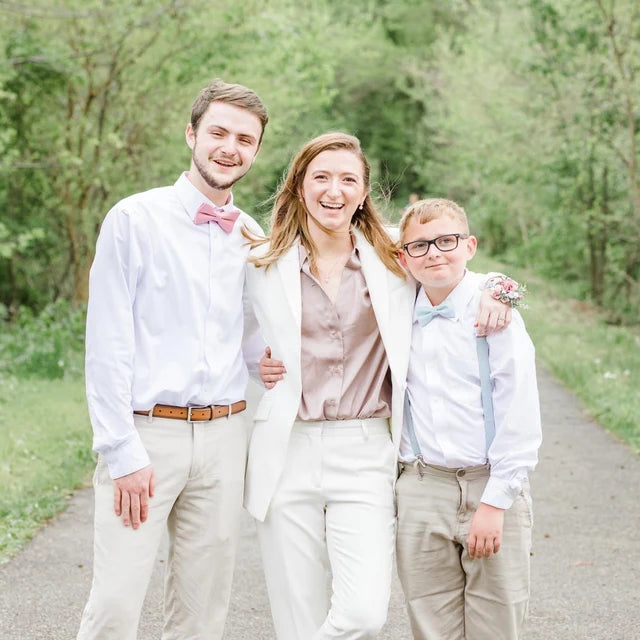 Family of three posing outdoors on a path with greenery. Boy wearing dusty blue bow tie and suspenders