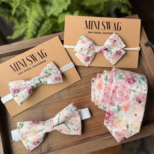 Floral bow ties and a necktie hair bow on Mini Swag packaging with a wooden surface and blurred greenery background.
