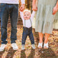 Child holding hands with adults in a forest setting wearing dusty sage suspenders