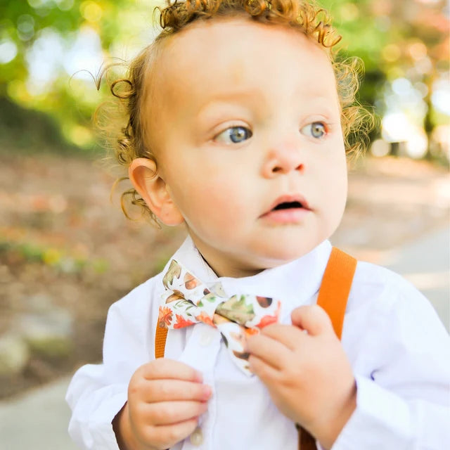 Child wearing a white shirt, floral bow tie, and orange suspenders outdoors.