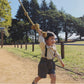 Child playing on a rope swing in a park with trees and grass in the  background wearing plum purple suspenders
