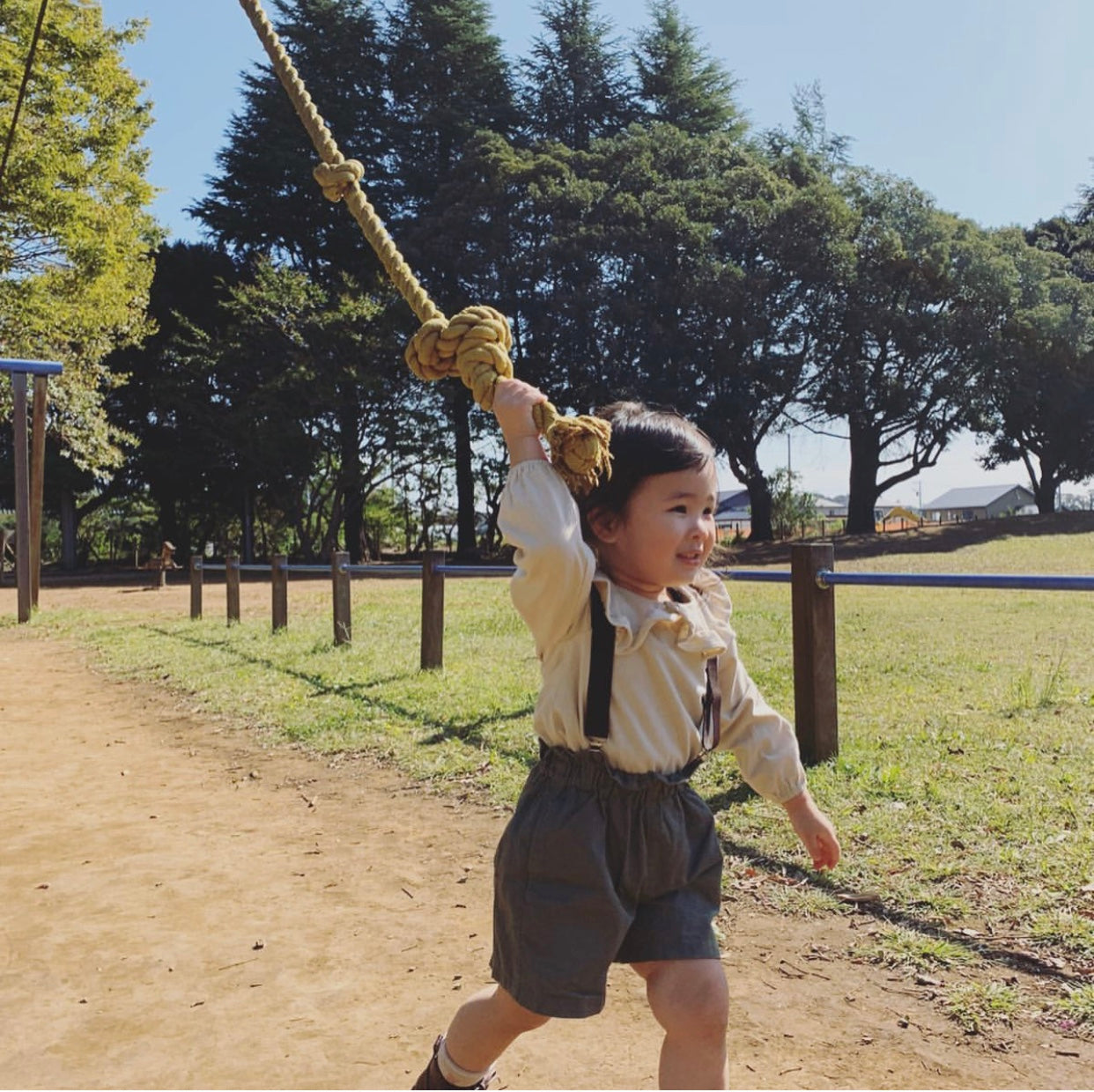 Child playing on a rope swing in a park with trees and grass in the  background wearing plum purple suspenders