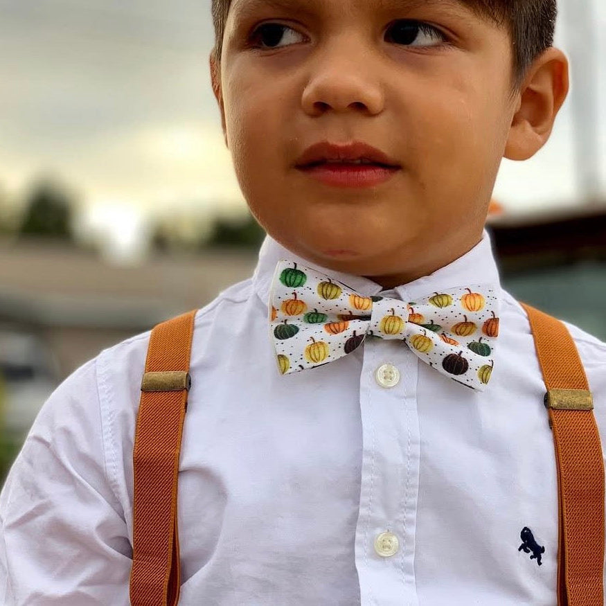 Young boy wearing a white shirt with a pumpkin patterned bow tie and brown suspenders.
