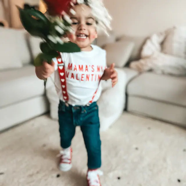 Child holding flowers with a 'Mama's My Valentine' shirt in a living room setting heart suspenders